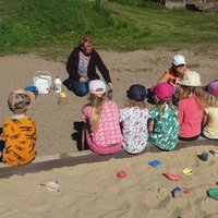 Lapset istuvat hiekkalaatikon reunalla ja kuuntelevat ja katselevat kertomusta Tyhmästä ja viisaasta miehestä.