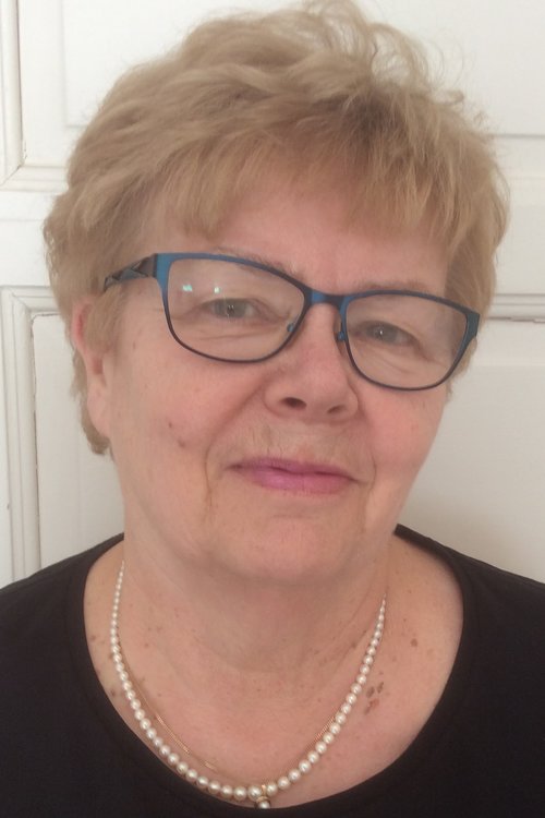 Hymyilevä vaaleatukkainen, silmälasipäinen nainen. Musta paita ja helmikaulanauha.