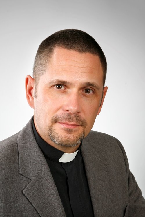 Lyhyt- ja tummatukkainen mies papinpaidassa.