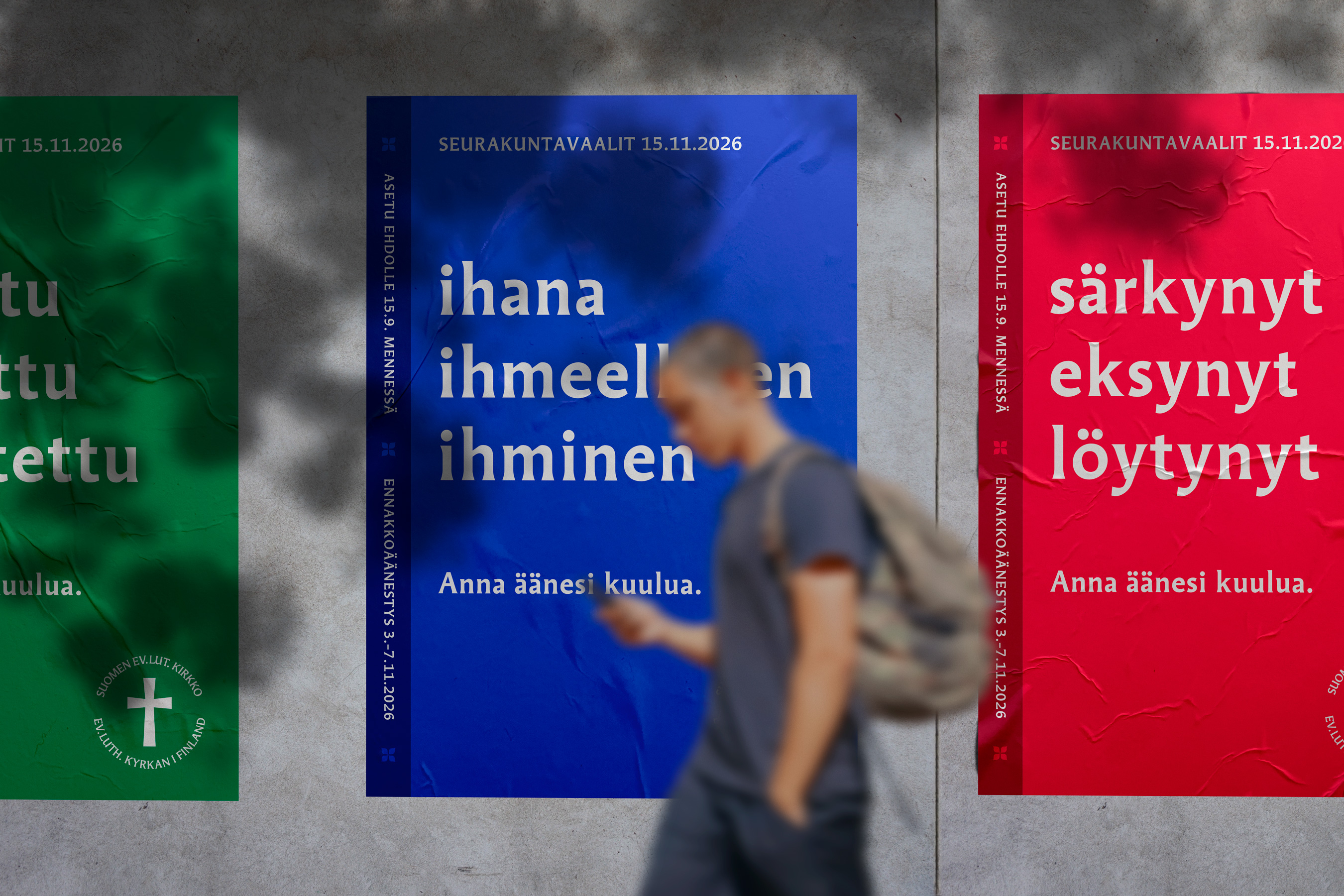 Kolme julistetta seinällä, joissa on eri värejä ja tekstejä. Henkilö kävelee ohi, taustalla näkyy punainen, si